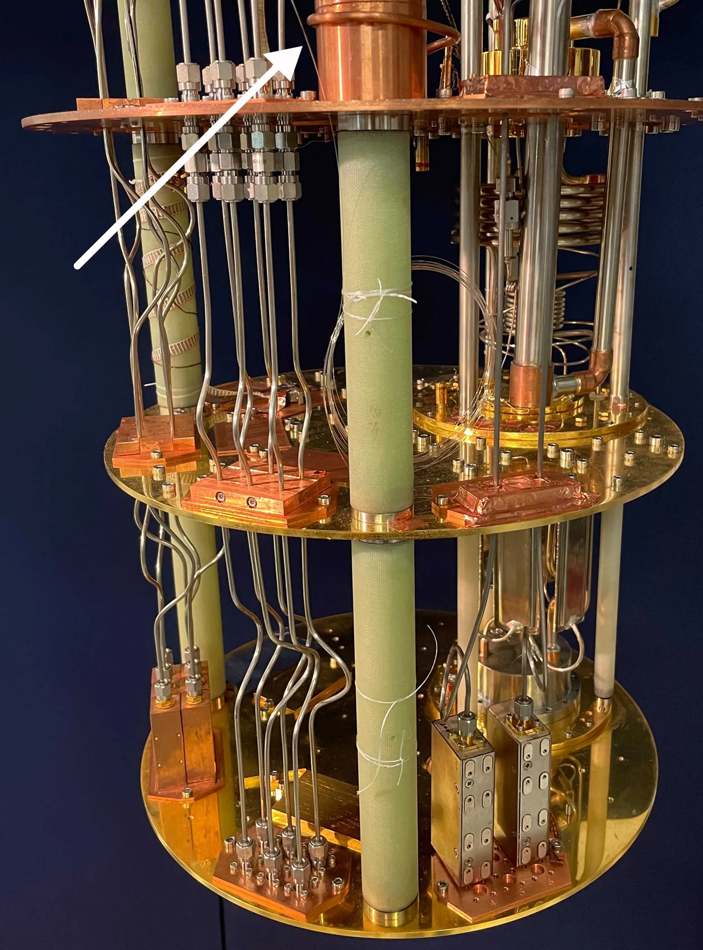 A gold and copper dilution-refrigerator assembly with many metal cables and a white arrow indicating an optical fiber used for superconducting-qubit control.