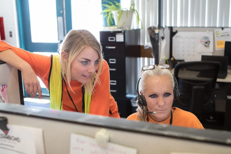 Two women in orange workwear review a computer at a call-center station, with one wearing a headset.