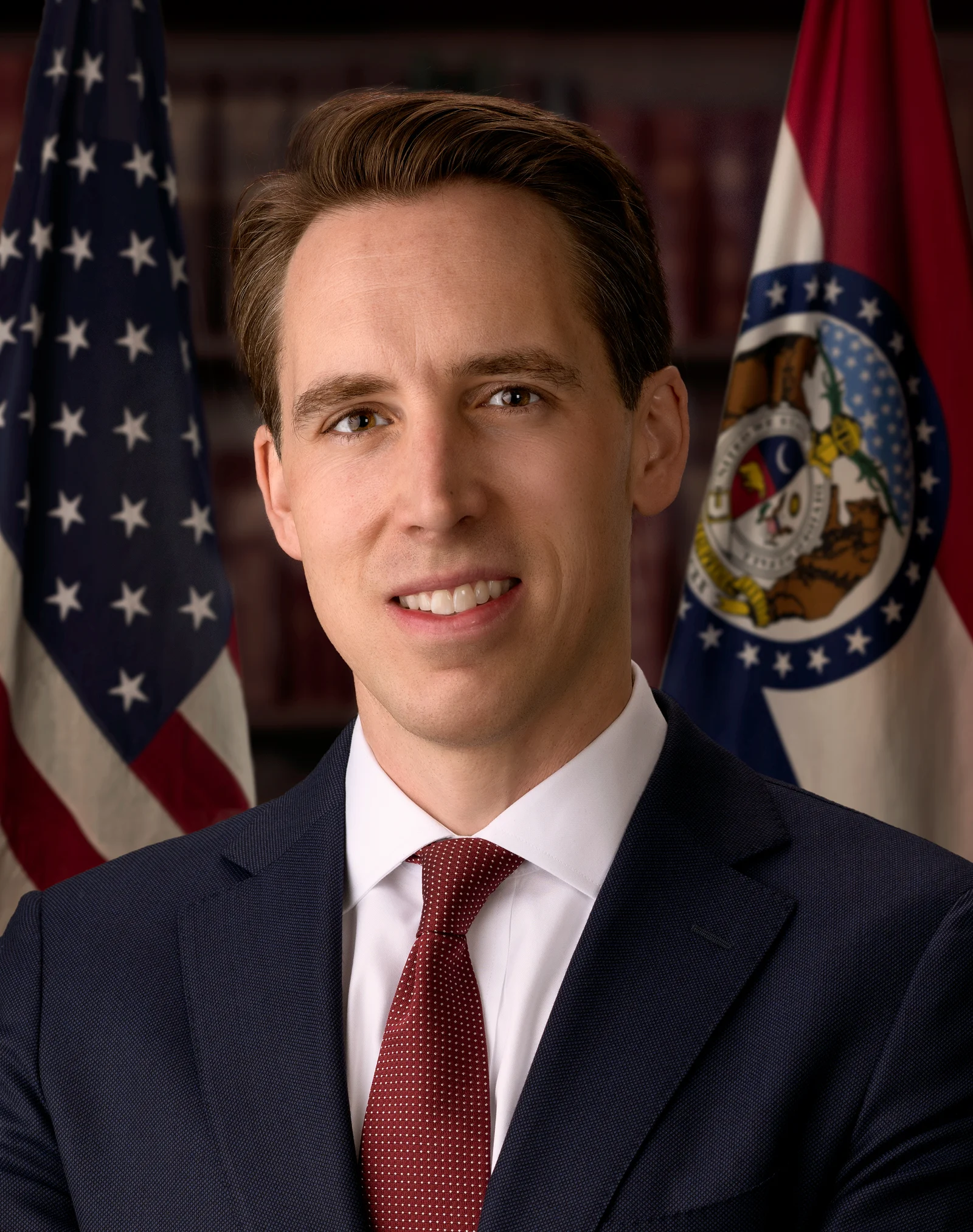 Josh Hawley poses in a dark suit and red tie between the United States and Missouri flags.