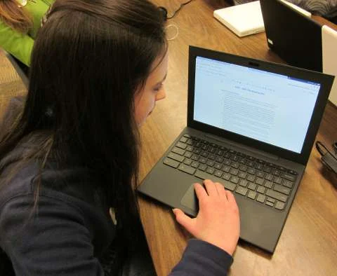 A student seated at a classroom table using a small laptop, viewed from above and behind